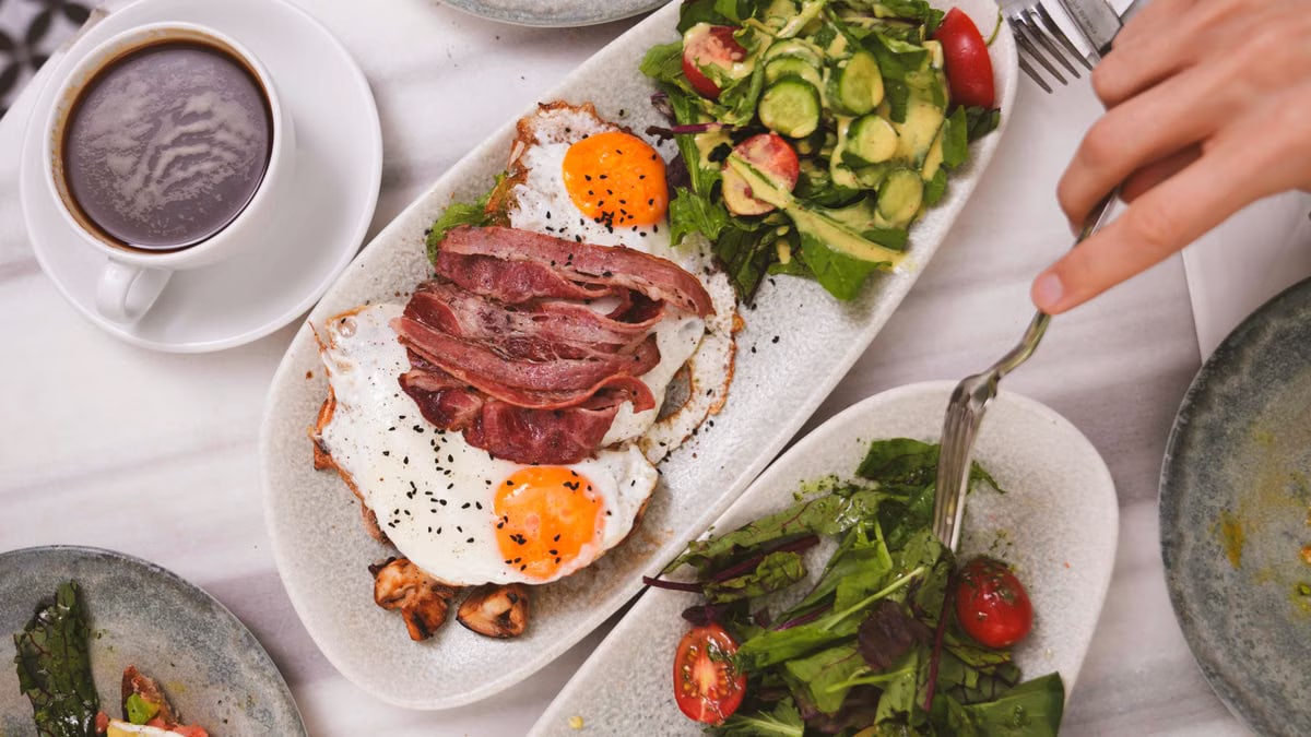 Top view of a brunch meal featuring fried eggs, bacon, fresh salad, and coffee.